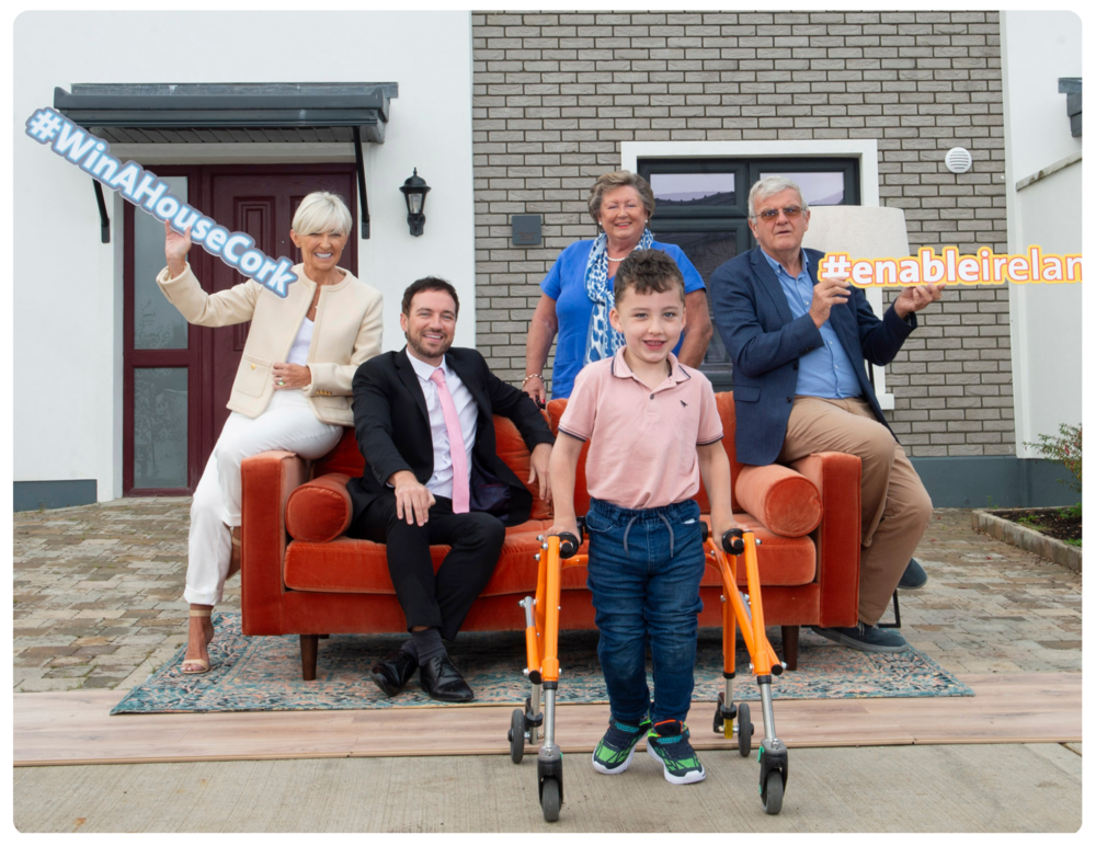 A photo taken outside the house to be won in Cork. Adam stands with his orange walking frame. Behind him a four people in formal clothing sat on an orange sofa. A woman in a cream blouse is sat on the left of the sofa holding a blue sign that says "#WinAHouseCork". A man in a blue shirt and suit jacket is sat on the right holding a sign that says "#EnableIreland".