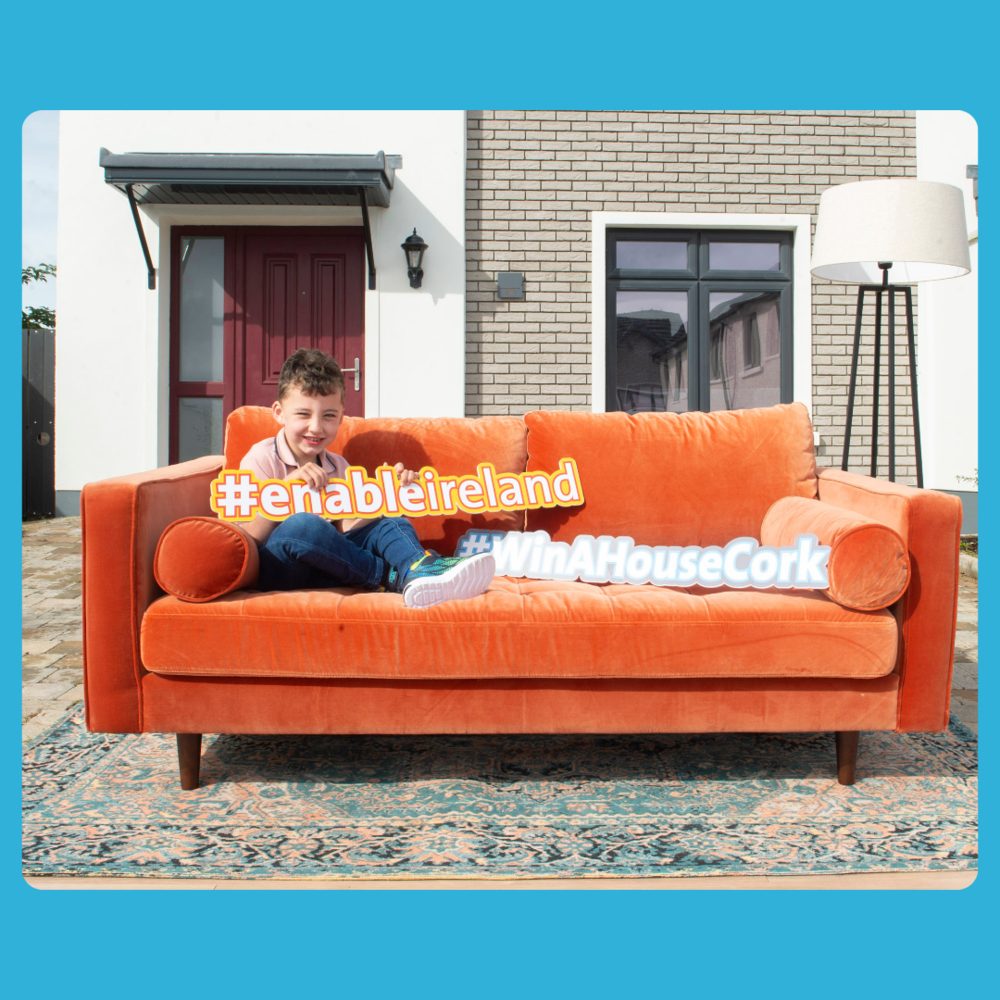 A photo taken outside the house to be won in Cork. Outside of the house is an orange sofa on which Adam sits holding an orange \